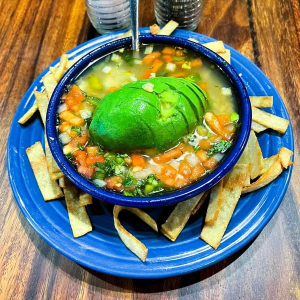 Mexican soup with sliced avocado, diced vegetables, and tortilla strips on a blue plate.