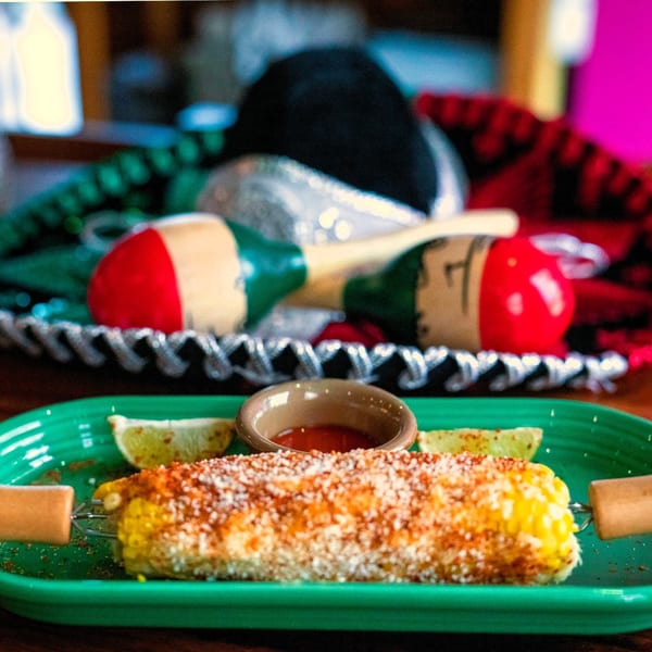 Elote, Mexican street corn, on a green plate with lime and sauce, maracas in background.