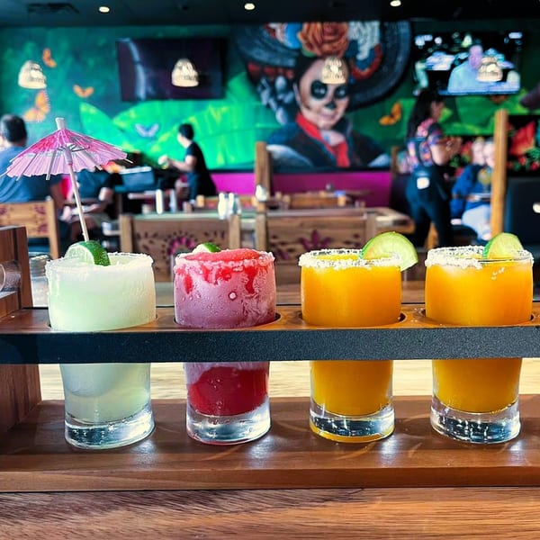 Four colorful margaritas in a flight board at La Patrona Cantina and Grill, with a vibrant mural in the background.