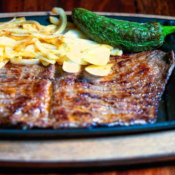 Grilled skirt steak with sautéed onions and a roasted green chili on a black plate.